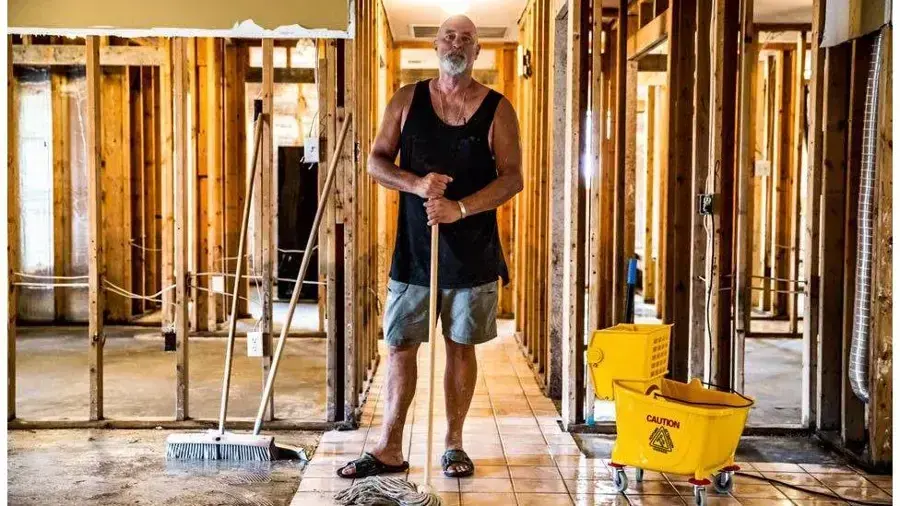 A homeowner with a mop and bucket in a hallway opened to the studs after a flood