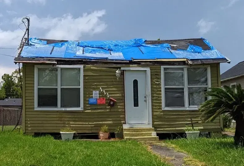 A small house with a blue tarp across its storm-damaged roof